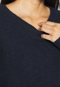 Granatowy sweter z dekoltem w serek wykonany z miękkiej wełny, o fakturowanej powierzchni i długich rękawach. Ręka spoczywa na kołnierzu dla podkreślenia detalu.