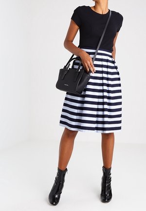 Frau trägt schwarzen Kurzarm-Top, knielangen dunkelblau-weißen horizontal gestreiften Rock, schwarze Knöchelstiefel und hält eine schwarze Umhängetasche.