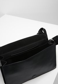 Černá kožená messenger taška s semišovým klopou, která má vnitřní kapsu a odnímatelný ramenní popruh pro všestrannost.