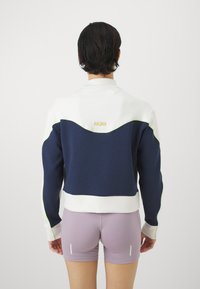 Marine- og hvit sweatshirt med buet design, som har grønne aksenter og "HOKA"-logo på baksiden. Glatt stoff, avslappet passform.