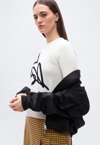 Γυναίκα με ίσια καστανά μαλλιά, που φοράει λευκό πουλόβερ με μαύρο σχέδιο, μουσταρδί καρό φούστα και μαύρο μπουφάν που έχει αποσπαστεί μερικώς.