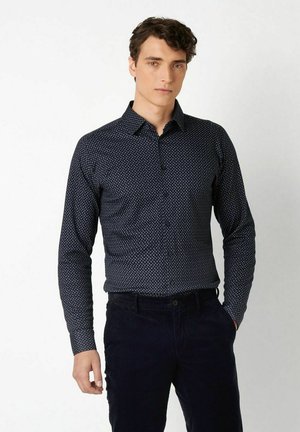 Junger Mann mit kurzen lockigen Haaren, der ein enganliegendes, dunkelblau gemustertes Langarmhemd und dunkle Hosen trägt, steht mit einer Hand in der Hosentasche.