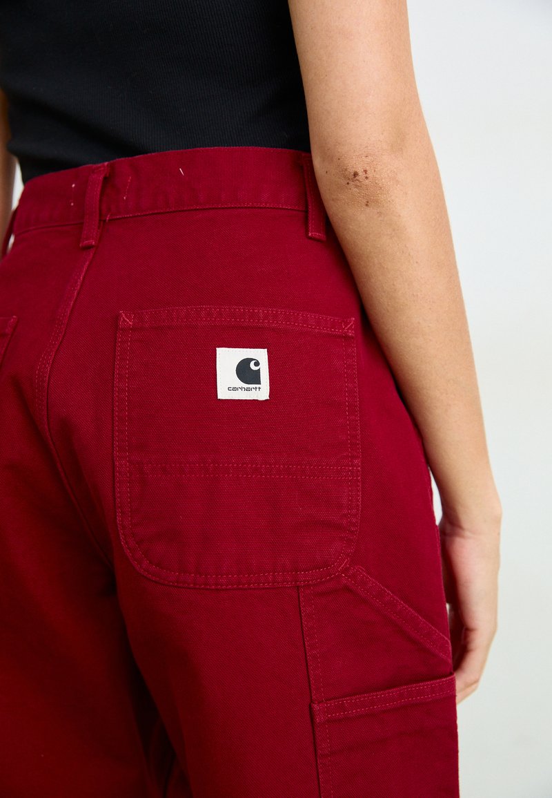 Bordeauxkleurige broek van stevig materiaal met een middelhoge taille, voorzien van achterzakken en een Carhartt logo-tag in een witte vierkant.