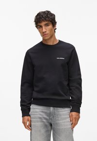 Sudadera negra hecha de tejido de forro polar, con cuello redondo, puños de punto y un pequeño logo blanco en el pecho. Combinada con unos vaqueros gris claro.