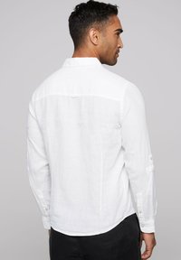 Camisa de lino blanca con detalle de botones en las mangas, cuello en ángulo y tela texturizada, mostrada desde atrás sobre un fondo neutro.
