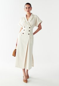 Kreemjas värvitoonis, lühikeste varrukatega nööpidest kleit, mis on varustatud vööga, külgaukudega ja kahe ees oleva taskuga, millel on ümmargused nupud.