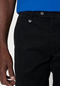 Pantaloni neri con chiusura a bottone e gancio metallico. Il tessuto è liscio, con una leggera texture opaca. Sono visibili piccole tasche.