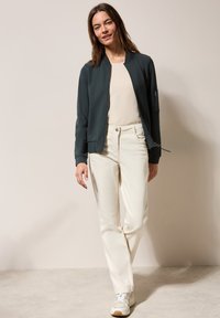 Giacca bomber verde scuro con polsini a costine, tasca con zip, indossata sopra un top beige e pantaloni bianchi. Tessuto leggero, vestibilità casual, abbinata a sneakers.