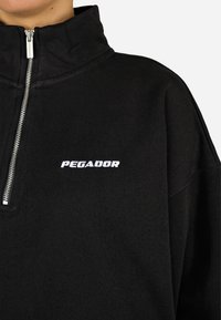 Melns puloveris ar augstu apkakli un sudraba rāvējslēdzēju, ar baltu "PEGADOR" logotipu priekšā. Mīksta, teksturēta auduma.