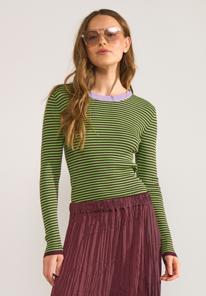 Femme portant un haut à manches longues rayé vert et noir, une jupe plissée bordeaux et de grandes lunettes de soleil teintées roses sur fond uni.