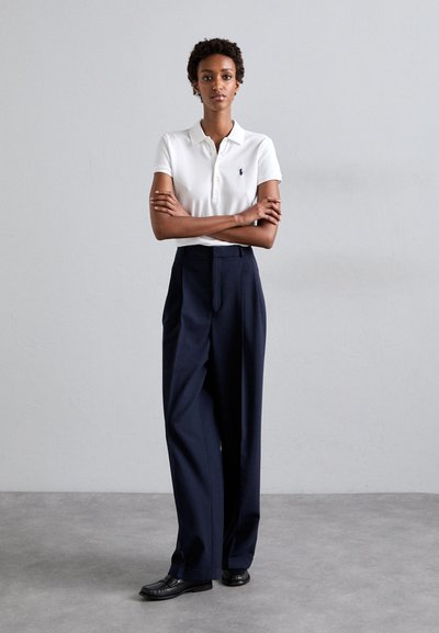 White polo shirt with button placket and embroidered logo, paired with high-waisted navy trousers and black shoes on a gray background.