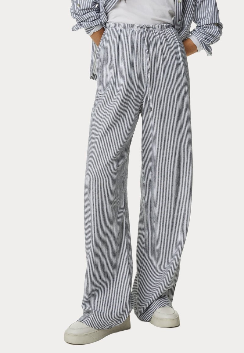 Personne portant un pantalon ample à jambes larges avec des rayures verticales bleu marine et blanches, une chemise rayée assortie, des baskets blanches et un t-shirt blanc rentré.