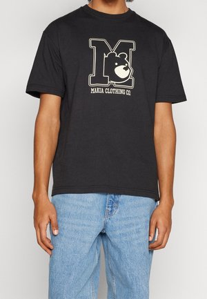 Svart bomullströja med en stor tryckt "M" och björnlåg i krämfärg. Kort ärm och rund halsdesign, kombinerad med blå denimjeans.