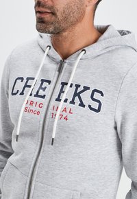Sudadera gris con cremallera fabricada en mezcla de algodón, con texto en azul marino y rojo, cordones y bolsillos frontales. Textura suave con un diseño casual.