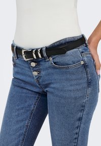 Femme portant un jean en denim bleu avec trois boutons argentés et une ceinture noire avec une boucle argentée, main posée sur la hanche.