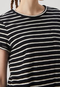 T-shirt à rayures noires et crème avec manches courtes, encolure ronde et petite poche sur le c ôté gauche. Texture en tissu de coton doux.