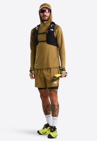 Sportowa odzież w odcieniach ziemi: bluzka z kapturem z długim rękawem i szorty. Czarna kamizelka nawadniająca i neonowożółte buty do biegania. Tatuaże widoczne na nogach.