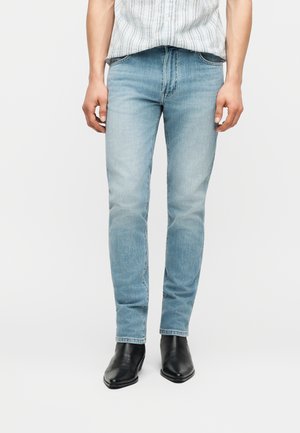 Bărbat purtând blugi slim fit albastru deschis și cizme negre cu vârf ascuțit, stând cu brațele relaxate pe lângă corp, pe un fundal simplu.