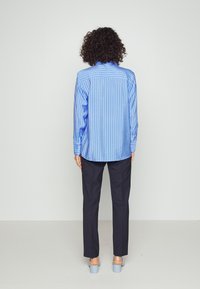 Blaue gestreifte Bluse mit langen Ärmeln, Knopfmanschetten und einem Rückenpasse, kombiniert mit dunklen Hosen und hellblauen Absatzschuhen. Rückansicht.
