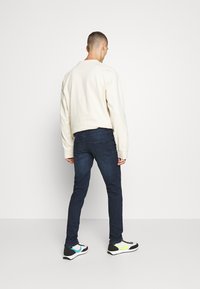 Herroutfit bestående av en krämfärgad sweatshirt, mörkblå slim-fit jeans och svarta sneakers med blå och gröna detaljer. Enkelt design, avslappnad stil.