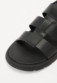 Sandale en cuir noir avec de larges sangles croisées, semelle plate et semelle intérieure texturée. Détails cousus le long des bords pour une durabilité renforcée.