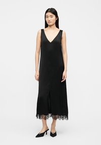 Femme portant une robe midi noire sans manches avec un col en V, une fente frontale, un ourlet en dentelle, et des talons noirs à bout pointu sur un fond blanc uni.