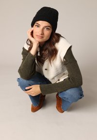 Gebreide zwarte beanie, crèmekleurige en groene gelaagde vest met knopen, groene trui mouwen, casual blauwe jeans en bruin suède enkellaarsjes.