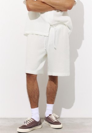 Homme portant un short blanc texturé, un haut blanc assorti, des chaussettes blanches et des baskets en toile marron, debout les bras croisés.