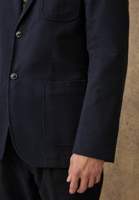 Navyblaues Jackett mit weicher Textur, ausgestattet mit zwei Knöpfen und einer aufgesetzten Tasche an der Seite, taillierte Passform mit klaren Linien.