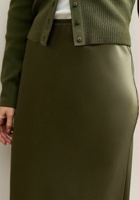 Gros plan sur une personne portant une jupe en cuir vert olive et un cardigan en tricot côtelé avec des boutons, le tout sur un haut blanc.