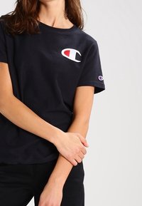 Camiseta de algodón negra con un logotipo bordado en blanco y rojo en el pecho y un pequeño logotipo en la manga. Mangas cortas, cuello redondo.