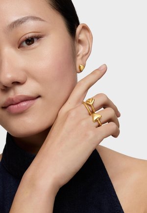 Donna che indossa un orecchino a forma di cuore dorato e tre anelli d'oro con design a cuore e freccia, che si tocca il viso con un dito.