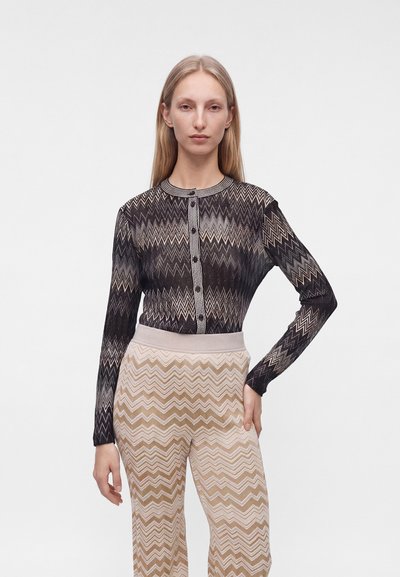 Femme debout, main sur la hanche, portant un haut boutonné à motif zigzag noir et beige et un pantalon assorti à motif zigzag, sur fond blanc.