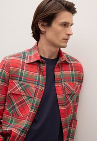 Camicia di flanella a quadri rossi e verdi con due tasche sul petto e bottoni in metallo, indossata sopra una t-shirt blu navy. Tessuto dalla trama liscia.