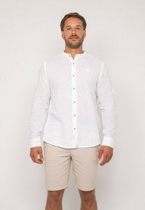 Homme portant une chemise blanche à col mandarin avec des boutons et un short beige, debout devant un fond clair uni.