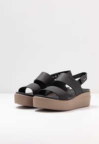 Schwarze Plateausandalen mit zwei breiten Riemen, verstellbarem Hinterriemen und einer hellbraunen Keilsohle. Glatte, matte Oberfläche mit strukturiertem Fußbett.