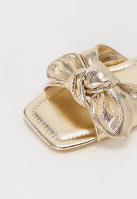 Guldfarget metallic sandaler med teksturert finish og en stor sløyfeakcent på stroppen. Rektangulær tå og flat såle design.
