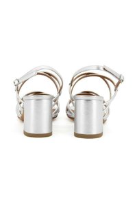 Sandali con tacco in argento dalla finitura metallica, con sottili cinturini incrociati e un robusto tacco a blocco, poggiati su una suola marrone.