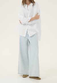 Person wearing an oversized white button-up shirt, light blue wide-leg jeans, and beige flat shoes, standing with arms crossed.