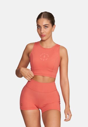 Kobieta w koralowym, bezrękawnym krótkim topie i dopasowanych szortach z wysokim stanem, pozująca na tle jednolitego tła, z ręką spoczywającą przy talii.