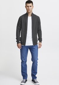 Cardigan zip grigio scuro con maniche lunghe sopra una t-shirt bianca, abbinato a jeans blu e sneakers nere. Design semplice e aderente.