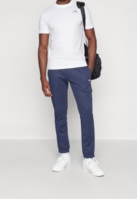 Bílý bavlněný tričko s malým černým logem, spárované s tmavě modrými cargo kalhotami s kapsami, a bílými teniskami. Černá taška přes rameno.