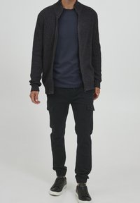 Cardigan zippé en gris foncé texturé sur un t-shirt bleu marine à col rond, associé à un pantalon cargo noir et des baskets noires avec des semelles blanches.