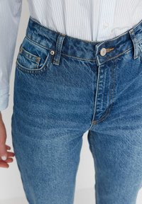 Blue denim jeans with a high waist, five-pocket design, and contrasting orange stitching. The fabric has a slightly faded texture.