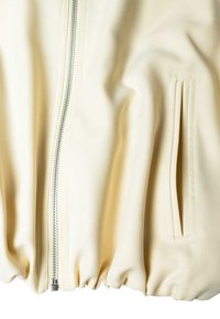 Veste en tissu crème vue de face montrant une fermeture éclair argentée et une poche fendue verticale avec contour cousu sur le côté droit.