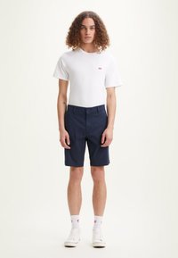T-shirt blanche à manches courtes avec un petit logo rouge, associé à un short bleu marine. Modèle portant des baskets blanches, accentuées de chaussettes blanches.