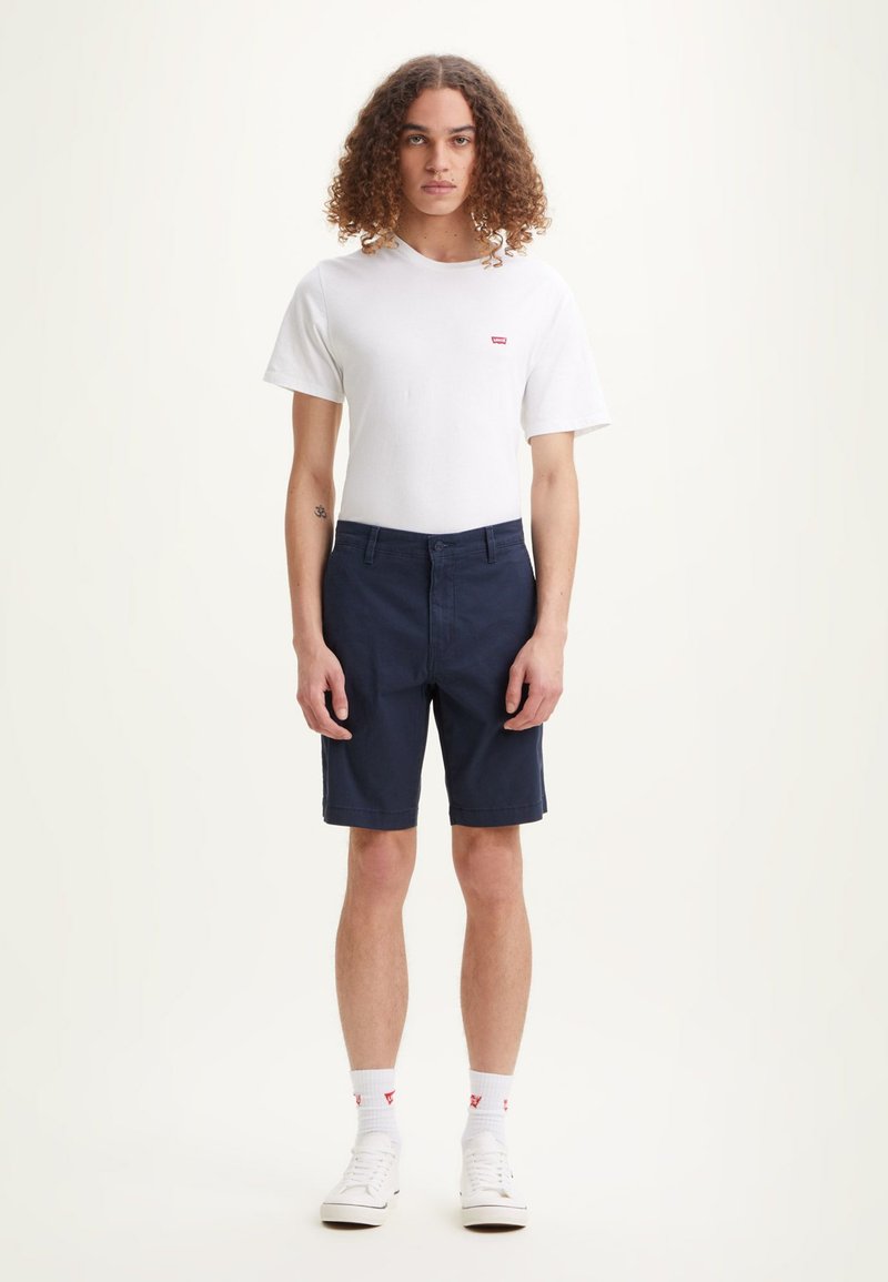 T-shirt blanche à manches courtes avec un petit logo rouge, associé à un short bleu marine. Modèle portant des baskets blanches, accentuées de chaussettes blanches.