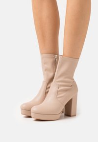 Beige nilkkuriset, jotka on valmistettu sileästä nahasta. Niissä on korkea lohkokorko, vetoketjusuljenta ja pehmeä, laskostettu muotoilu.