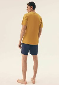Koszulka z krókim rękawem w kolorze musztardowym z okrągłym dekoltem, zestawiona z granatowymi szortami. Materiał wygląda na lekki i gładki. Stojąc boso.