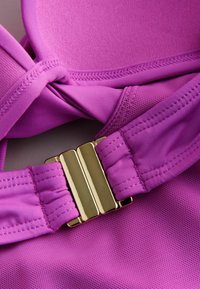 Top sportivo rosa con coppe sagomate, tessuto testurizzato e un fermaglio dorato per la regolazione. Sono visibili spalline lisce e cuciture.